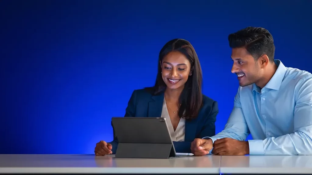 Two colleagues smiling at a tablet on a desk, blue studio background
