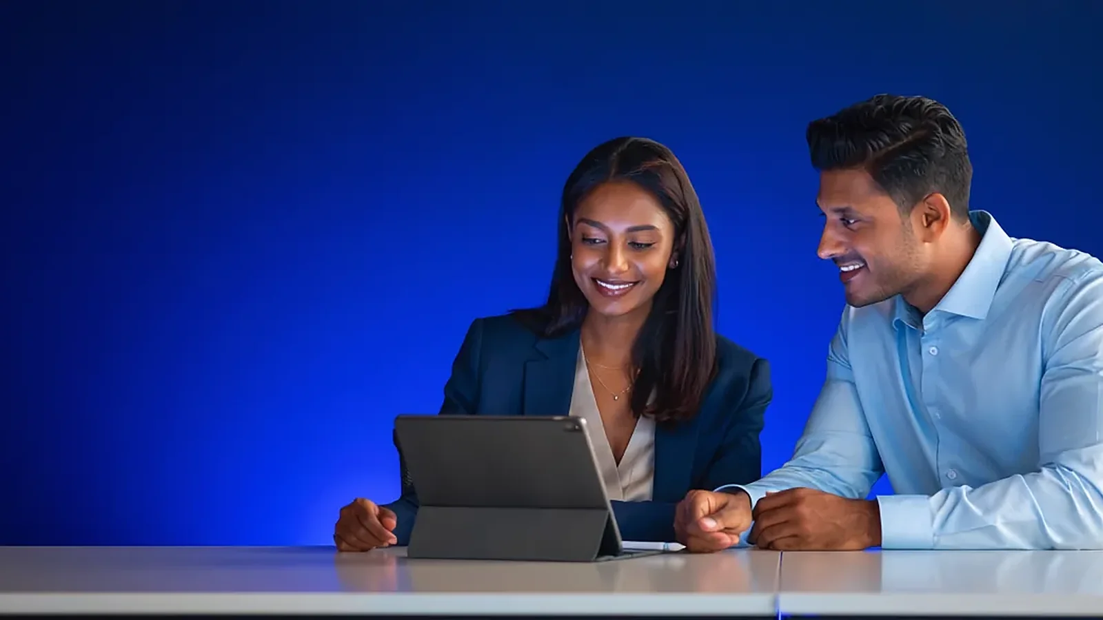 Two colleagues smiling at a tablet on a desk, blue studio background